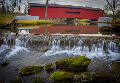 A red covered bridge spans a calm river, with a waterfall and mossy rocks in the foreground, surrounded by trees.
