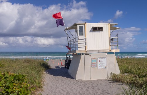 Lifeguard tower on a beach with red and purple flags, surrounded by sand and grass, under a blue sky with clouds.