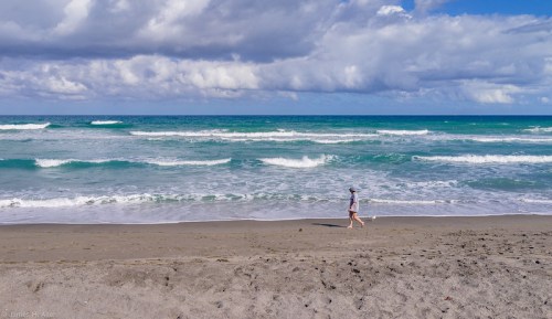 A person walks along a sandy beach with waves crashing and clouds in the sky.