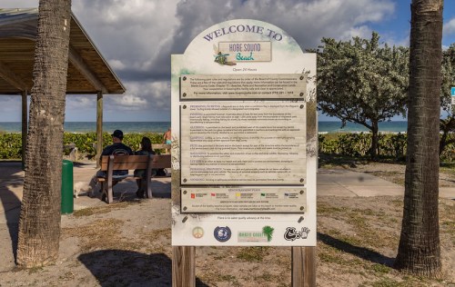 A welcome sign for Hobe Sound Beach with beach guidelines, overlooking the ocean and two people sitting on a bench.