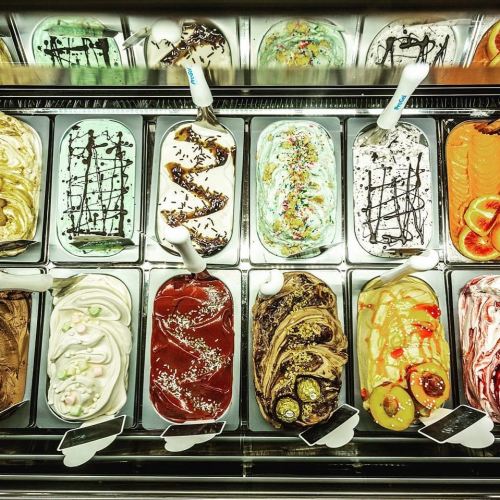 A variety of colorful gelato flavors displayed in metal containers, with toppings and sauces visible.