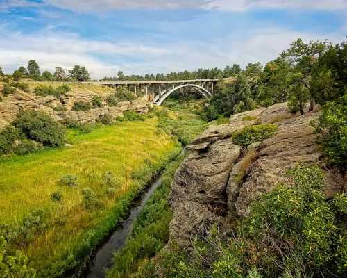 A scenic view of a bridge spanning a lush green valley with rocky terrain and a winding stream below.