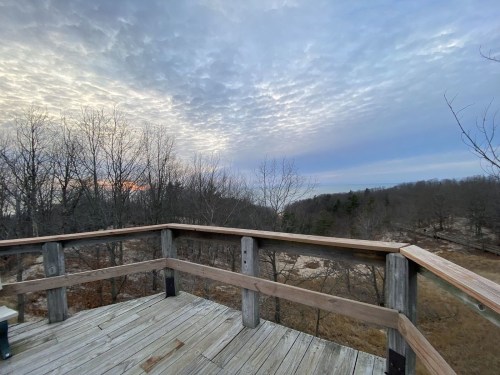 A wooden deck overlooks a landscape of bare trees and rolling hills under a cloudy sky at dusk.