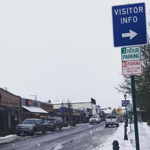 A snowy street scene with a "Visitor Info" sign and parking regulations, featuring shops and parked cars.