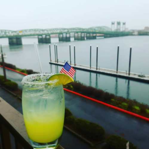 A refreshing cocktail with a lime and small American flag, overlooking a calm river and bridge on a cloudy day.