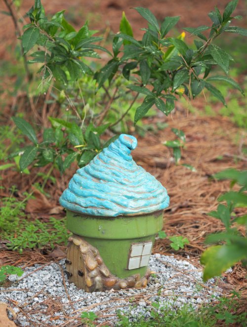 A whimsical plant pot shaped like an ice cream cone, with a blue swirl on top, surrounded by greenery and gravel.
