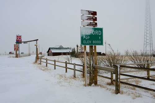 Snow-covered landscape with a sign for Buford, elevation 8000, and directional signs near a rustic building.