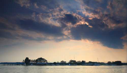 A serene waterfront scene with dark clouds and a hint of sunlight peeking through, reflecting on the water's surface.