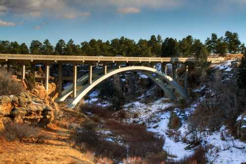 A modern arch bridge spans a rocky canyon, surrounded by trees and patches of snow under a blue sky.