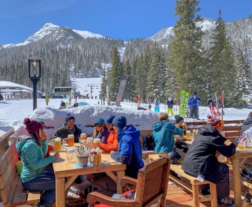 A group of people enjoying drinks at an outdoor table in a snowy mountain setting, with skiers in the background.