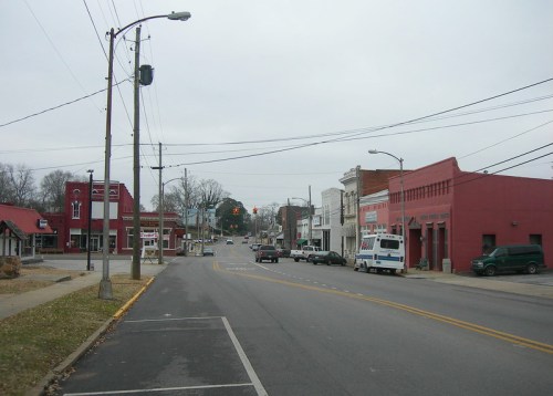 A quiet small town street with shops, parked cars, and overcast skies. Power lines run along the road.