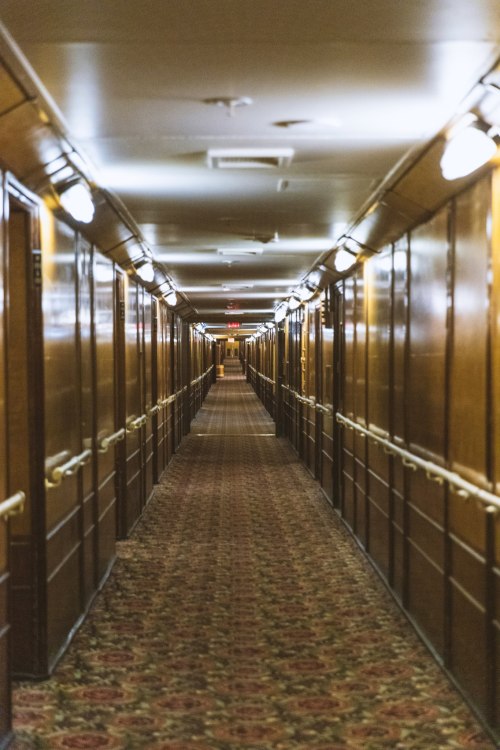 A long, dimly lit hotel corridor with wooden walls and patterned carpet, lined with doors on either side.
