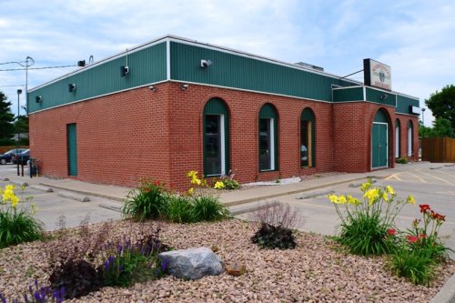 A brick building with green accents, surrounded by flower beds and a parking lot under a cloudy sky.