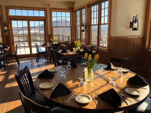 Bright restaurant interior with wooden tables, chairs, and large windows showcasing a scenic view. Fresh flowers on tables.