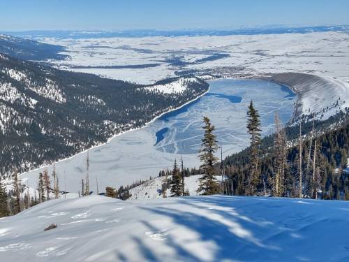 A snowy landscape with a frozen lake surrounded by evergreen trees and distant mountains under a clear blue sky.
