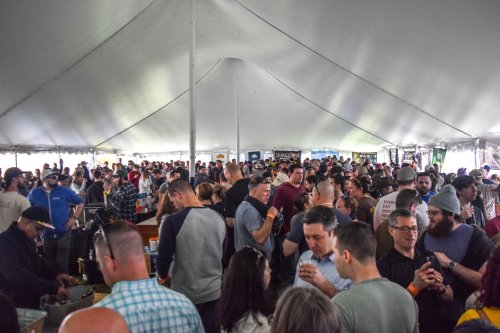 A large crowd of people socializing under a tent at an outdoor event, with food and drinks visible.
