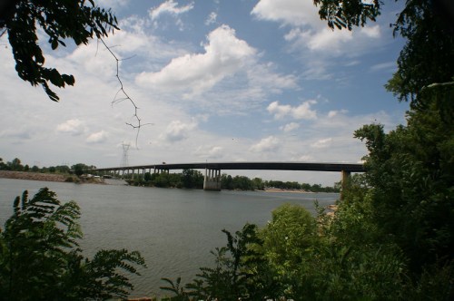A bridge spans a river under a partly cloudy sky, surrounded by greenery and trees on the riverbank.