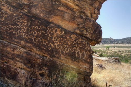 Petroglyphs etched into a rocky surface, featuring wavy, zigzag patterns against a backdrop of grassy terrain and hills.