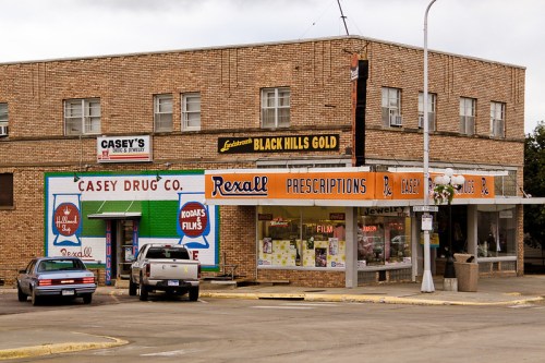 A vintage drugstore with colorful signage, parked cars, and a brick building in a small town setting.
