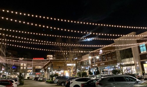 A nighttime view of a shopping area with string lights overhead and parked cars in the foreground.