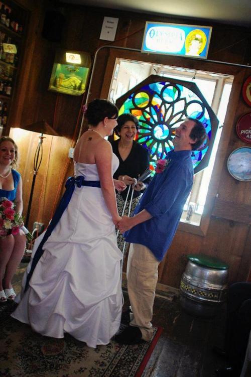 A couple exchanges vows in a cozy room, surrounded by friends, with a colorful stained glass window in the background.