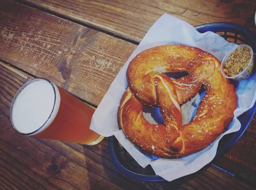 A large, golden-brown pretzel with coarse salt, served with mustard, alongside a glass of pale beer on a wooden table.