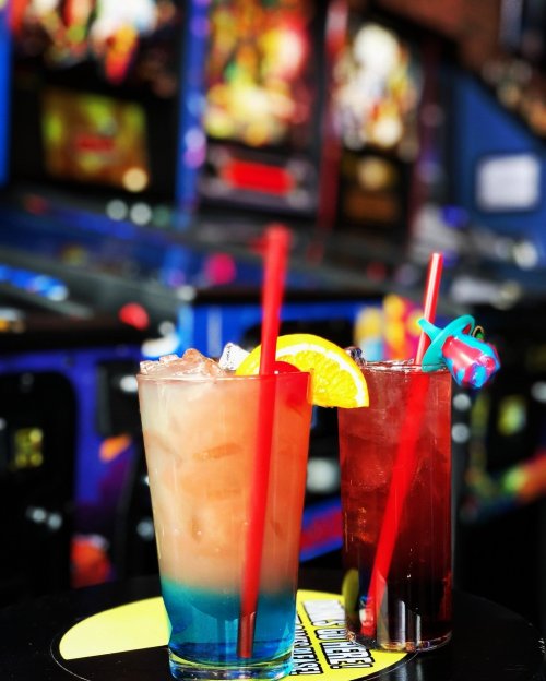 Two colorful cocktails with straws, garnished with orange slices, in front of a vibrant arcade game backdrop.