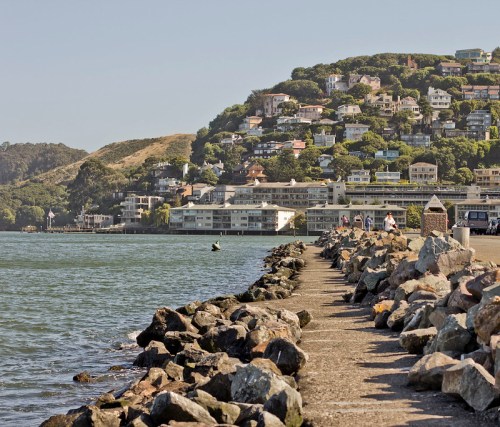 A rocky pier extends into the water, with hillside homes and greenery in the background under a clear sky.
