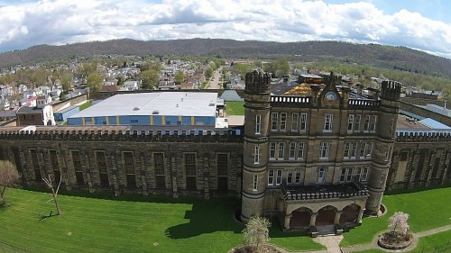 Aerial view of a historic stone building with towers, surrounded by green grass and a small town in the background.