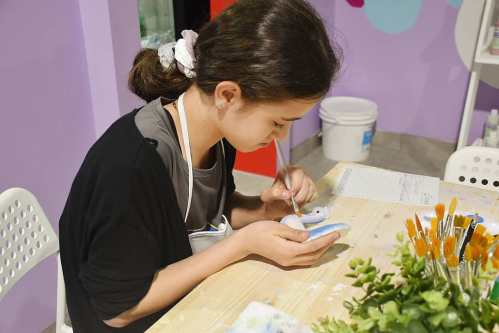 A young girl sits at a table, painting a small object with a brush in a colorful, creative space.