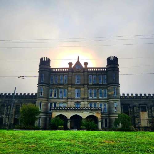 Historic stone building with towers, set against a cloudy sky and sun peeking through, surrounded by green grass.