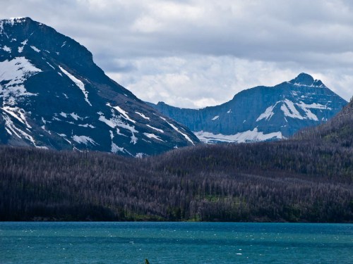 A scenic view of mountains with snow-capped peaks, overlooking a calm blue lake under a cloudy sky.