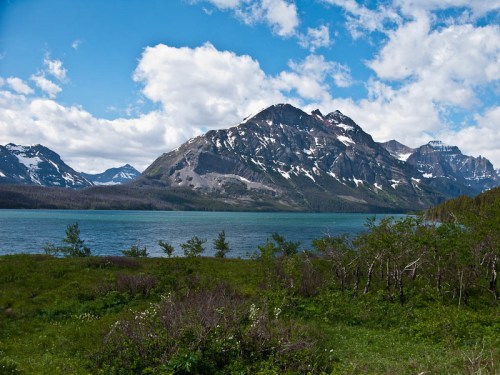 A scenic view of a lake surrounded by mountains, with lush greenery in the foreground and a blue sky with clouds.