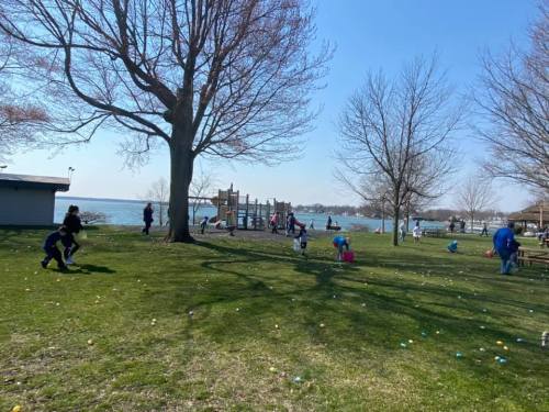 A sunny park scene with children hunting for eggs on grass, a playground in the background, and a lake visible.