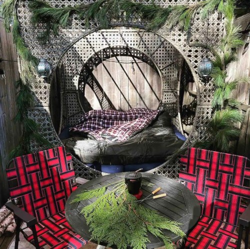 Cozy outdoor nook with a circular bed, surrounded by greenery, and two red plaid chairs at a small table.