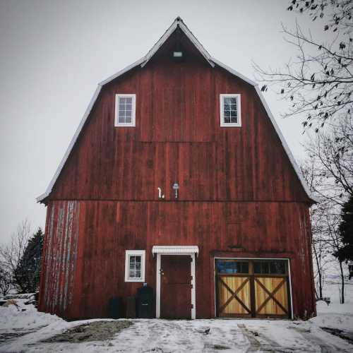 A red barn with a peaked roof, surrounded by snow and bare trees, under a gray winter sky.