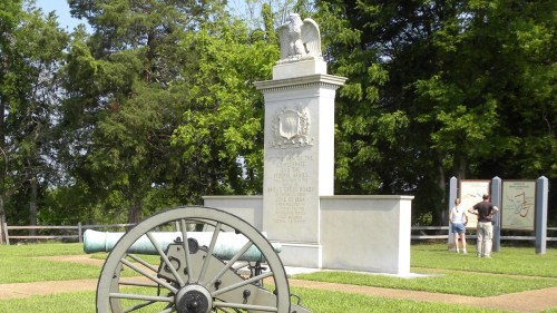 A historic monument with an eagle sculpture, surrounded by greenery and a cannon in the foreground. Visitors are nearby.
