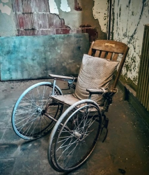 An old, wooden wheelchair with a striped pillow, set against a peeling wall in a dimly lit room.