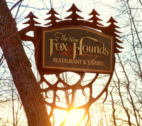 Sign for "The New Fox & Hounds Restaurant & Tavern" with a forest silhouette, illuminated by sunset.