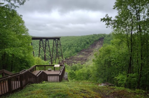 A scenic view of a green valley with a wooden overlook and an old trestle bridge in the background under cloudy skies.