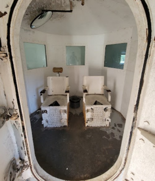 Interior of an old, empty room with two white chairs and small windows, showing signs of wear and neglect.