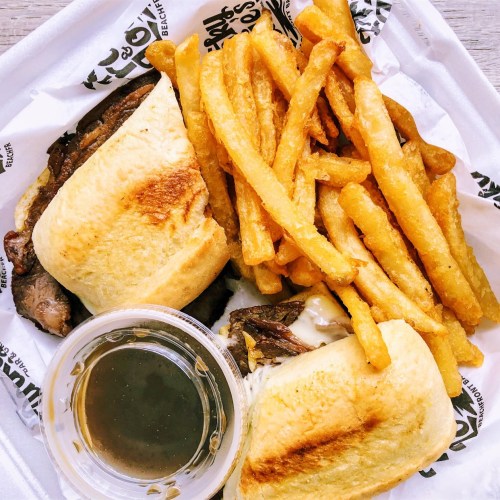 A plate featuring a sandwich with meat and melted cheese, accompanied by crispy French fries and a small cup of sauce.