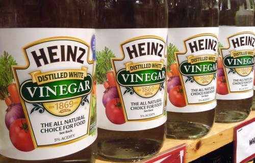 Four bottles of Heinz distilled white vinegar lined up on a shelf, featuring colorful labels with vegetables.