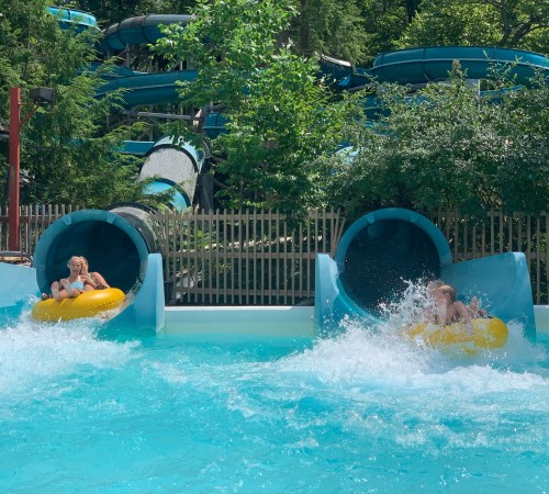 Two children slide down water slides into a pool, splashing water around them, surrounded by greenery.