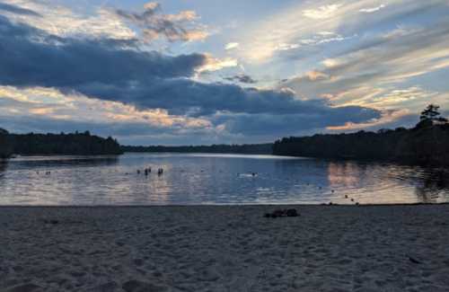 A serene lake at sunset, with clouds reflecting in the water and silhouettes of people swimming in the distance.