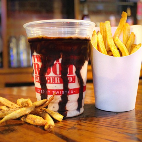 A cup of chocolate dessert with a drizzle, next to a container of golden fries on a wooden table.