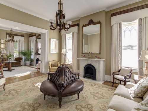 Elegant living room with a brown tufted sofa, ornate chandelier, and large windows, featuring classic decor and a fireplace.