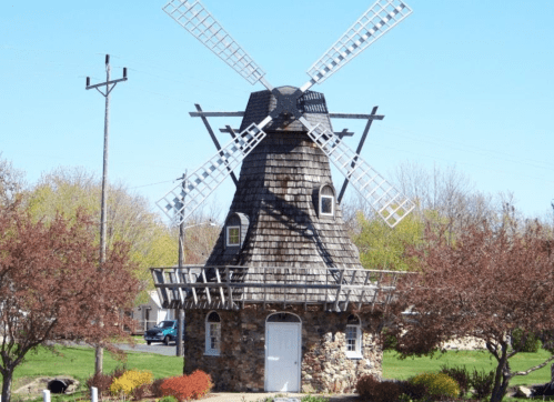 A stone windmill with a shingled roof and large wooden blades, surrounded by greenery and trees under a clear blue sky.