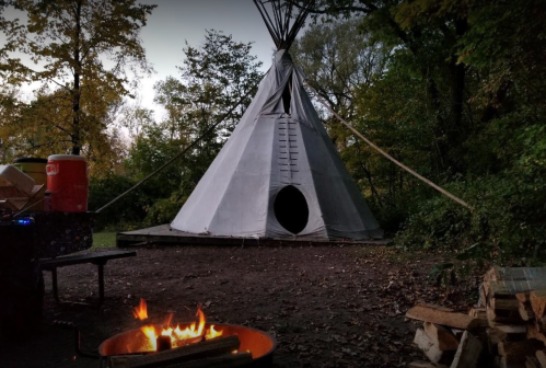 A teepee in a wooded area with a campfire in the foreground, surrounded by autumn foliage.