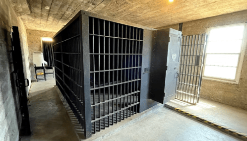 A view of a dimly lit jail cell with metal bars and an open door, featuring a concrete floor and walls.